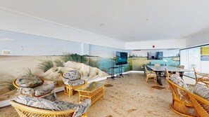 Ein Zimmer mit Wandgemälde zum Strandthema, Korbmöbeln, einem Flachbildfernseher und einem Esstisch mit Stühlen.