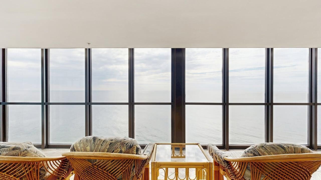 A room with large windows, wicker furniture, and a glass table.