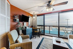A room with a television, a ceiling fan, a large window with a view of the sea, a sofa, a table, and a chair. - Convenient studio right on the beach with pools & W/D (Ocean City)