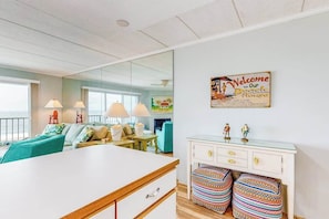 A modern living room with a sofa, a coffee table, and a dining area with a white cabinet and colorful ottomans. - Direct oceanfront top floor condo w/outdoor pool, balcony, easy beach access (Ocean City)