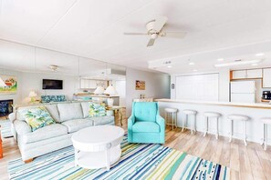 A modern living room with a sofa, a round coffee table, a teal armchair, and a kitchen area with a bar. - Direct oceanfront top floor condo w/outdoor pool, balcony, easy beach access (Ocean City)