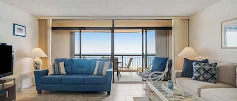 A living room with a blue sofa, a white sofa, a glass coffee table, a TV, and a view of the sea through large windows.