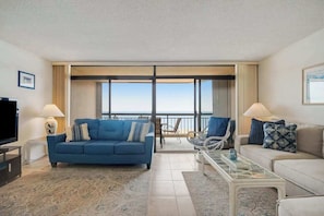 A living room with a blue sofa, a white sofa, a glass coffee table, a TV, and a view of the sea through large windows. - Fountainhead Towers 802 (Ocean City)