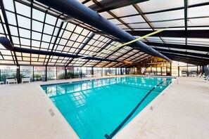 Ein Innenpool mit Glasdach, klarem Wasser und weißen Liegestühlen rundherum.