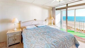 Un dormitorio con cama, dos mesitas de noche con lámparas, un ventilador de techo y un ventanal con vistas a la playa.
