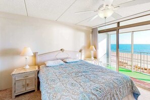 Ein Schlafzimmer mit einem Bett, zwei Nachttischen mit Lampen, einem Deckenventilator und einem großen Fenster mit Blick auf den Strand.