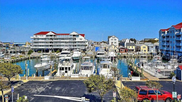 A marina with boats docked, a large building with a red roof, and a red car parked in the foreground. - Emerson Towers 301 (Ocean City)