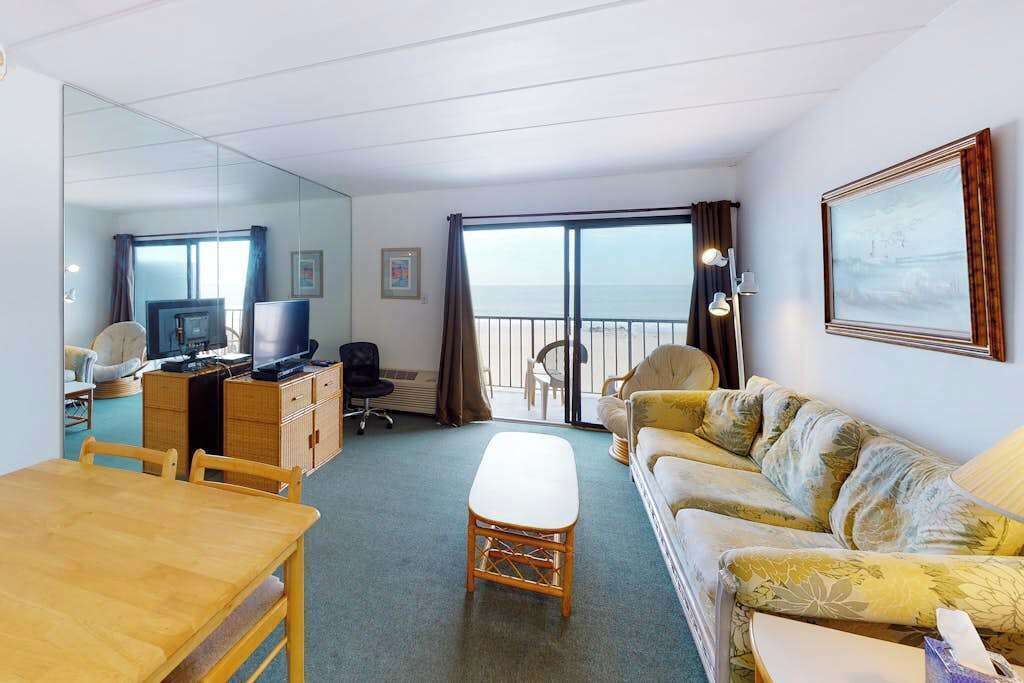 A hotel room with a balcony, a dining area, a sofa, a TV, and a desk.