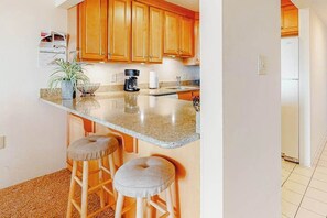 A kitchen with wooden cabinets, a granite countertop, two stools, a coffee maker, and a refrigerator. - Vacation Retreat in Oceanfront Complex w/Free WiFi, Central AC, W/D (Ocean City)