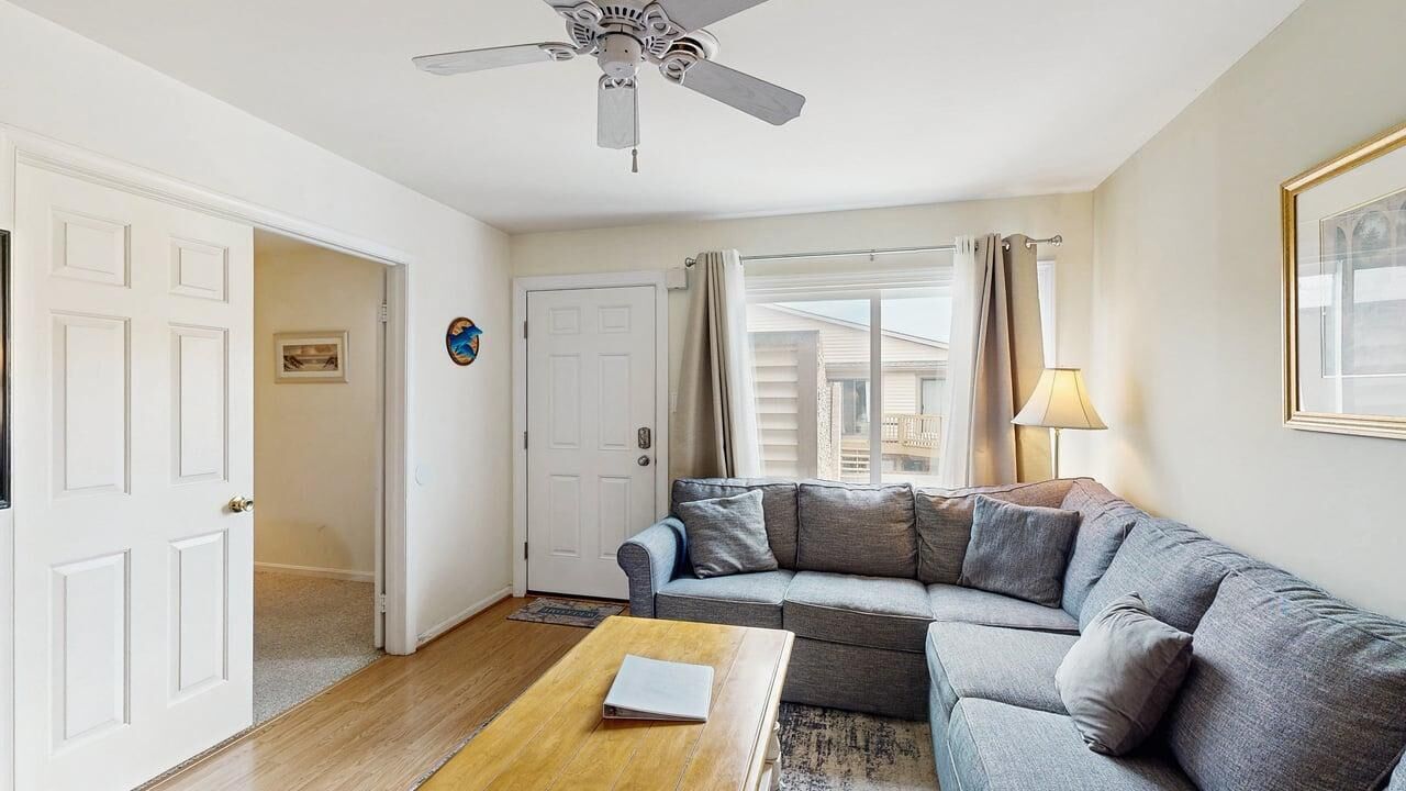 Una sala de estar con un sofá gris, una mesa de centro de madera, un ventilador de techo y una puerta que da a otra habitación.