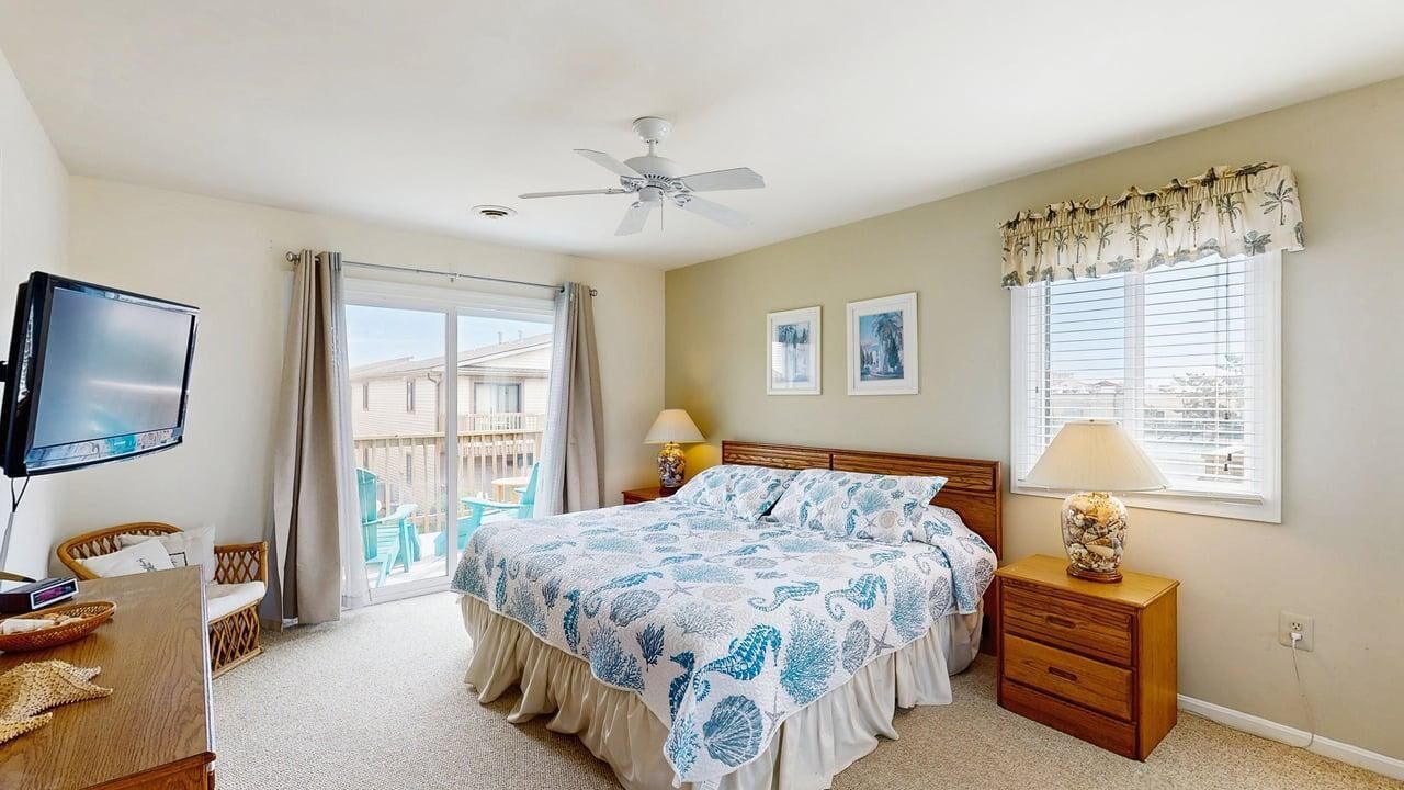 A bedroom with a bed, a TV, a ceiling fan, a window with curtains, and a balcony view.