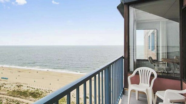 A balcony with a view of the beach, white plastic chairs, and a glass door leading to an interior space. - Unique Condo w/Side Views of the Beach, Private Balcony, Shared Pools, Gym, A/C (Ocean City)