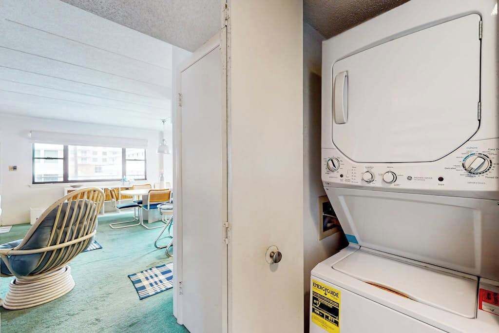 A laundry room with a washing machine, a chair, and a door leading to a room with a window and a table.