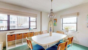 Una habitación con una mesa blanca, sillas azules, cestas de mimbre, una tabla de surf y una lámpara colgante.