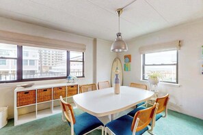 Ein Zimmer mit einem weißen Tisch, blauen Stühlen, Korbwannen, einem Surfbrett und einer hängenden Lampe.