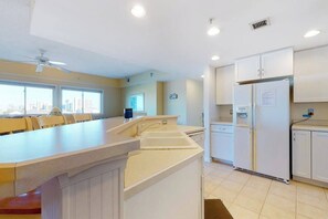 A kitchen with a central island, white cabinetry, a refrigerator, and a view of the city through large windows. - Charming Bayfront Condo Close to the Beach w/Shared Pool, Free WiFi, Private W/D (Ocean City)