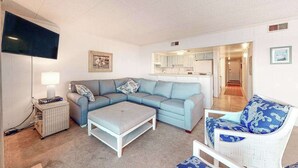 Een woonkamer met een hoekbank, een witte fauteuil, een bijzettafeltje en een televisie.