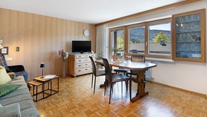 Una sala de estar moderna con piso de madera, un comedor con mesa y sillas, un televisor de pantalla plana y un ventanal con vistas al exterior.