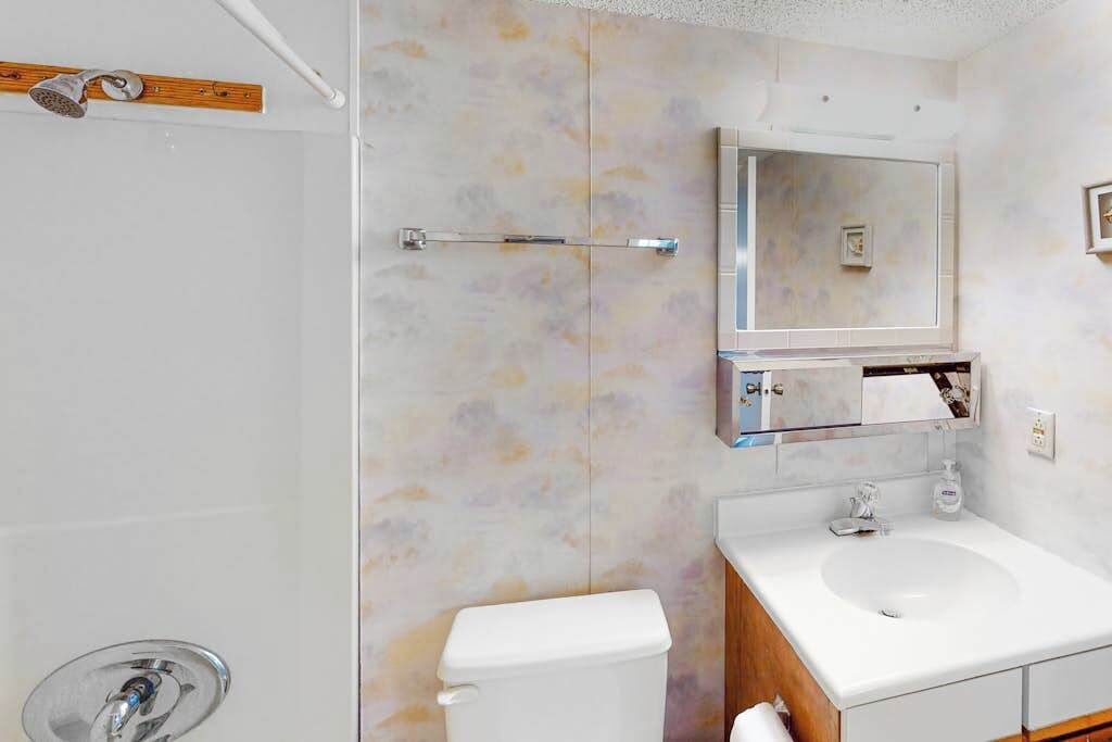 A bathroom with a toilet, sink, mirror, shower, and a wooden shelf.