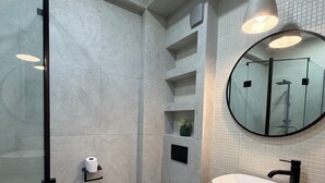 A modern bathroom with a glass shower enclosure, a toilet, a sink with a round mirror, and built-in shelves. - Cozy minimalistic apartment in Bagebi (T'bilisi)