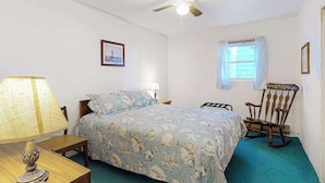 Une chambre à coucher comprenant un lit, un bureau, une chaise, un fauteuil à bascule, un ventilateur de plafond, une fenêtre avec des rideaux et un tableau encadré accroché au mur.