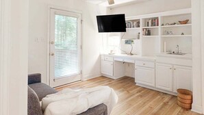 Un espace de vie compact comprenant un canapé, un téléviseur à écran plat fixé au mur, une kitchenette avec des meubles blancs et un plancher en bois.