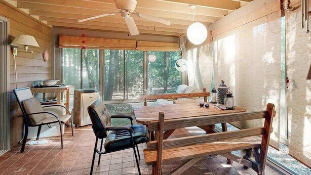 Une table à manger en bois, des chaises, un ventilateur de plafond et une grande fenêtre donnant sur les arbres.