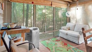 Une pièce avec une grande fenêtre, un canapé blanc, une table avec un bol et des papiers, une chaise et un radiateur portatif.