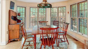 Une salle à manger avec une table ronde, six chaises en bois, un piano et de grandes fenêtres donnant sur un paysage arboré.