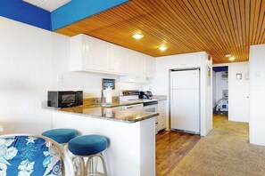 A compact kitchen with white cabinetry, a microwave, a stove, and a refrigerator. There is a small island with stools. - Beachloft 2H (Ocean City)