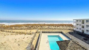 Une piscine avec vue sur la plage et l’océan, entourée d’une clôture et d’un bâtiment.