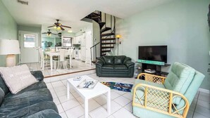A living room with a sofa, a coffee table, a TV, a wicker chair, a staircase, and a kitchen in the background. - Canal View Townhouse w/Private Deck, Washer/Dryer, & Private Balcony (Ocean City)
