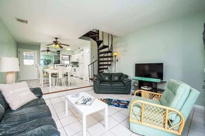 Ein Wohnzimmer mit einer Couch, einem Couchtisch, einem Fernseher, einem Korbstuhl, einer Treppe und einer Küche im Hintergrund.