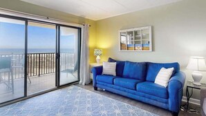 Una habitación con un sofá azul, un ventanal de vidrio que da a un balcón, una mesita con una lámpara y un cuadro en la pared.