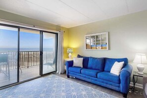 Ein Zimmer mit blauer Couch, einer Glastür, die auf einen Balkon führt, einem kleinen Tisch mit Lampe und einem Bild an der Wand.