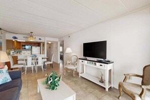 Een woonkamer met een televisie, een zithoek met stoelen, een keuken op de achtergrond, en een witte console tafel.
