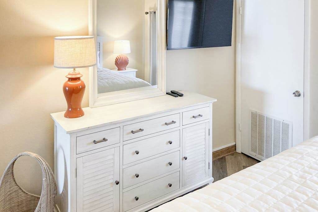 Une commode blanche avec des tiroirs, un miroir, une lampe et un téléviseur accroché au mur.