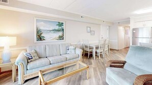 A living room with a sofa, a coffee table, a dining table with chairs, and a kitchen area with cabinets. - Marigot Beach 1206 (Ocean City)