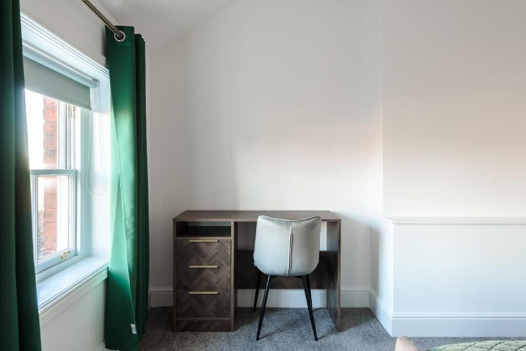 Quarto de canto com escrivaninha, cadeira e janela com cortinas verdes.