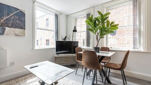 A modern living room with a TV, dining table, chairs, a plant, and large windows.