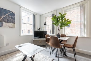 Een moderne woonkamer met een televisie, een eettafel, stoelen, een plant en grote ramen.