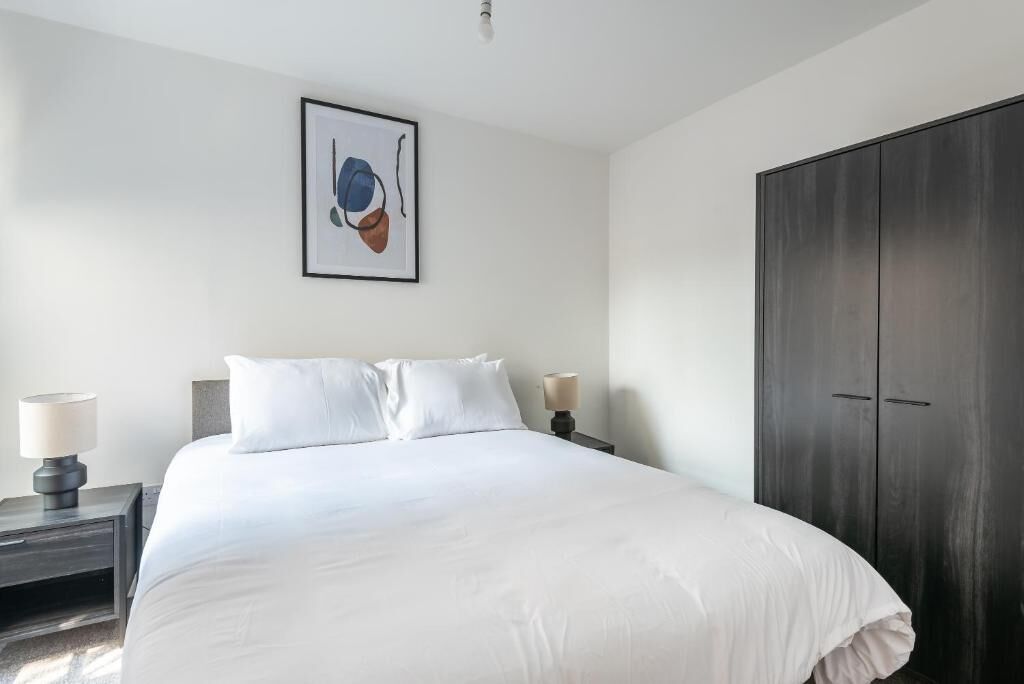 Une chambre à coucher avec un lit, deux tables de chevet avec des lampes, une armoire sombre et un tableau encadré au mur.