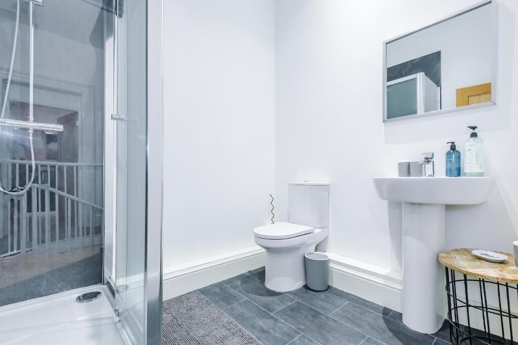 Un baño moderno con cabina de ducha de vidrio, inodoro blanco, lavamanos pedestal y una pequeña mesa redonda con una taza.