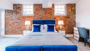 A bedroom with a brick wall, a bed with blue bedding, two bedside tables with lamps, and a desk with a chair.