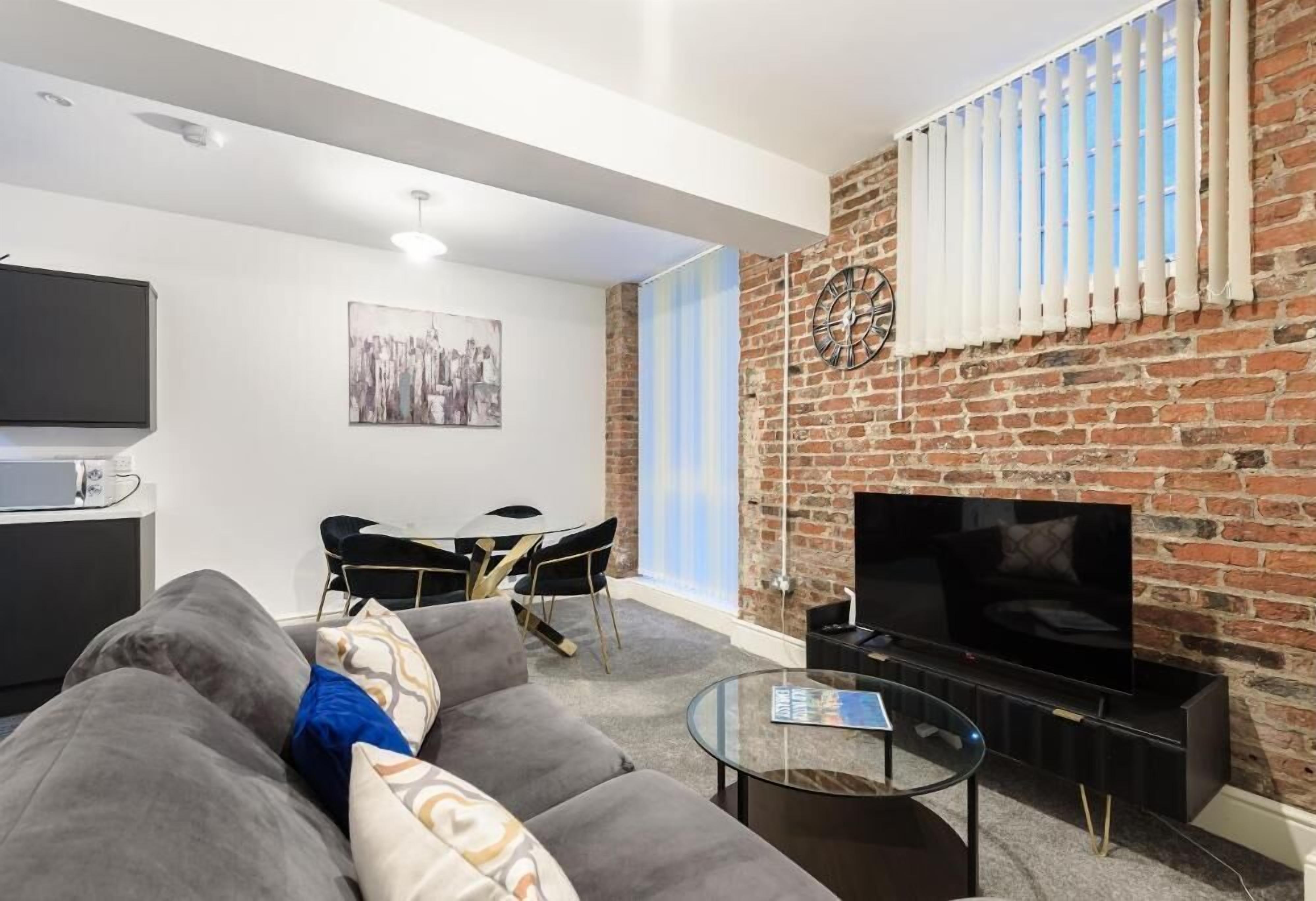 A modern living room with a brick wall, a flat-screen TV, a sofa, a coffee table, and a dining area with chairs and a table.