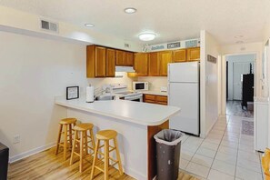 A kitchen with wooden cabinets, a white island with stools, a refrigerator, and a microwave. - Lovely bayside condo with wading pool, balcony, beach access, W/D, & central AC (Ocean City)