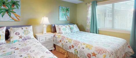 A bedroom with two beds, a nightstand, a lamp, and two paintings of palm trees.