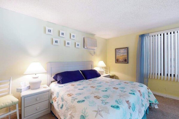 A bedroom with a bed, two bedside tables with lamps, a chair, a wall-mounted air conditioner, and a window with blinds. - Beachfront condo with balcony, W/D, & outdoor pool (Ocean City)