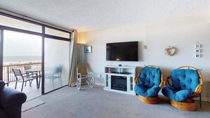 Quarto com uma grande porta de vidro deslizante que dá para uma varanda, televisão de tela plana na parede, duas cadeiras de vime com almofadas azuis e um pequeno móvel branco para entretenimento.