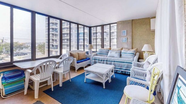 Uma sala de estar moderna com grandes janelas, um conjunto de sofá, móveis de vime branco e um tapete azul.